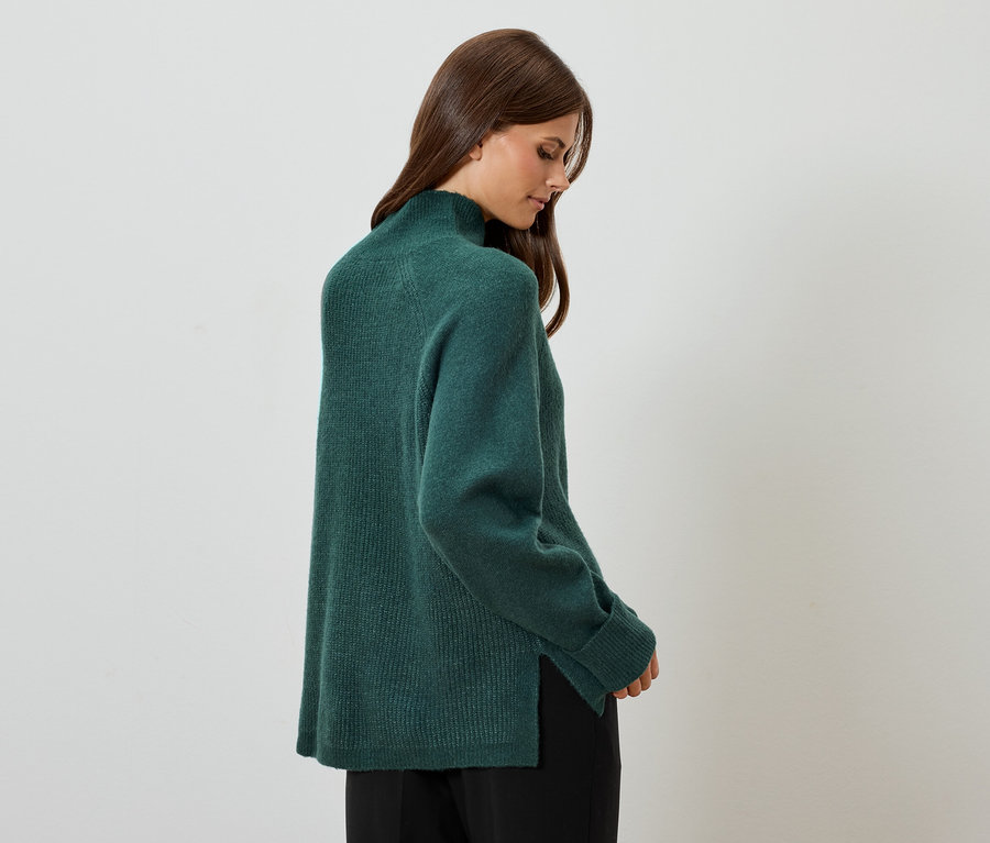 Kadın yeşil bir kazak ve siyah pantolonla poz veriyor.