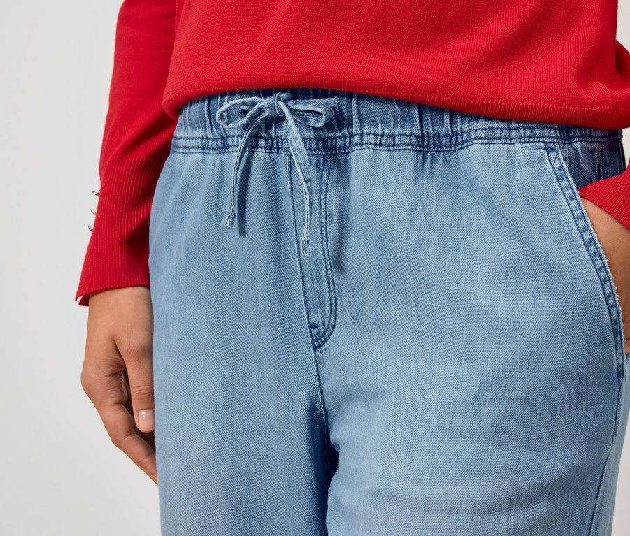 Kırmızı bir kazak ve açık mavi kot pantolon giyen bir kişinin kısmi görünümü, eli cebinde.