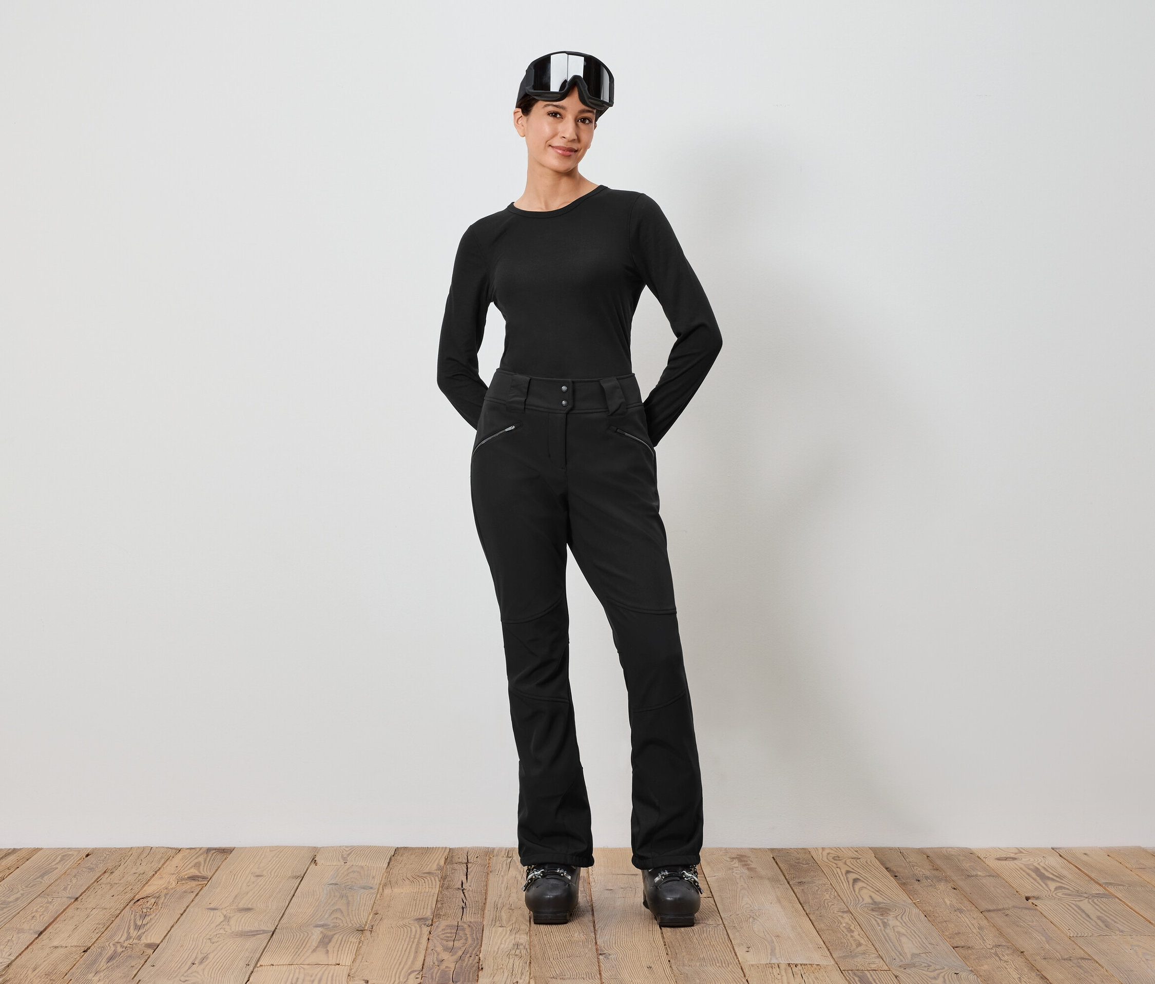 Başında kayak gözlüğü olan siyah kayak kıyafetli bir kadın beyaz bir duvarın önünde duruyor.