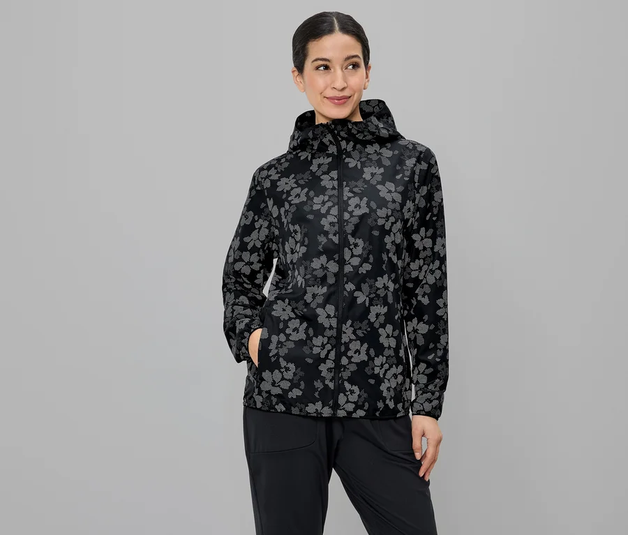 Gri bir arka plan önünde kapüşonlu, çiçek desenli bir ceket giyen ve siyah pantolonunun cebine elini sokan bir kadın.