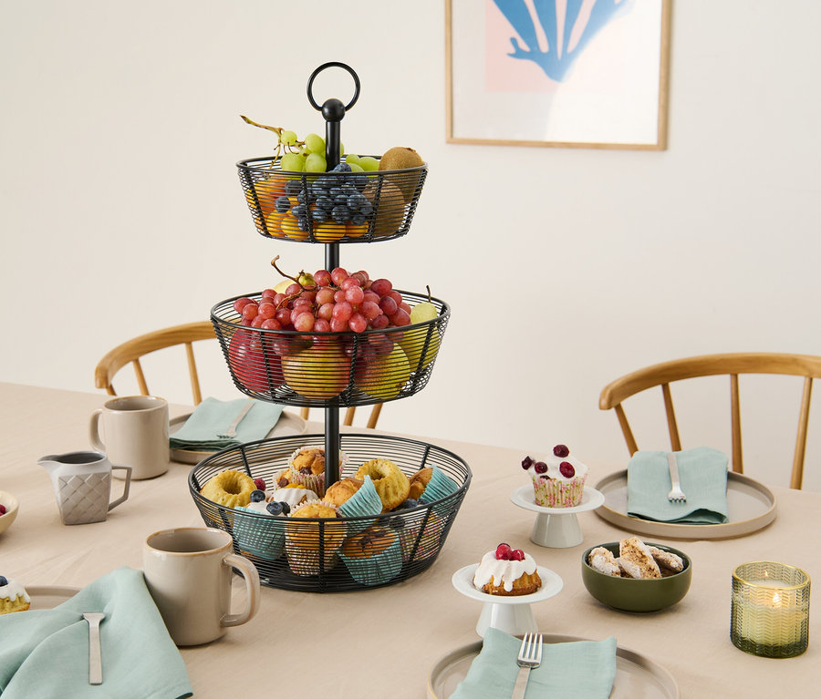 Metal telden yapılmış üç katlı bir meyve sepeti, masa üzerinde hamur işleri, meyveler ve tabaklarla birlikte duruyor.