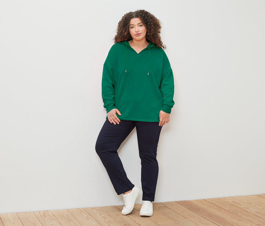 Kıvırcık saçlı bir kadın, yeşil kapüşonlu sweatshirt, lacivert pantolon ve beyaz spor ayakkabı giyerek beyaz bir duvarın önünde poz veriyor.