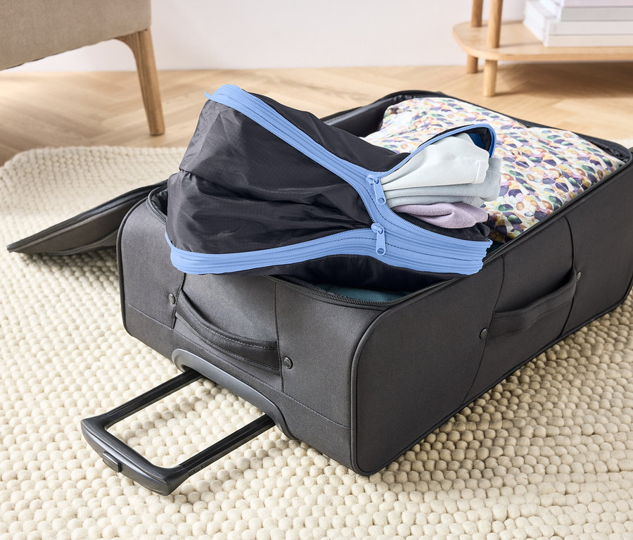 Halı üzerinde açık, gri bavul, giysilerle ve mavi fermuarlı siyah bir ambalaj küpüyle dolu.