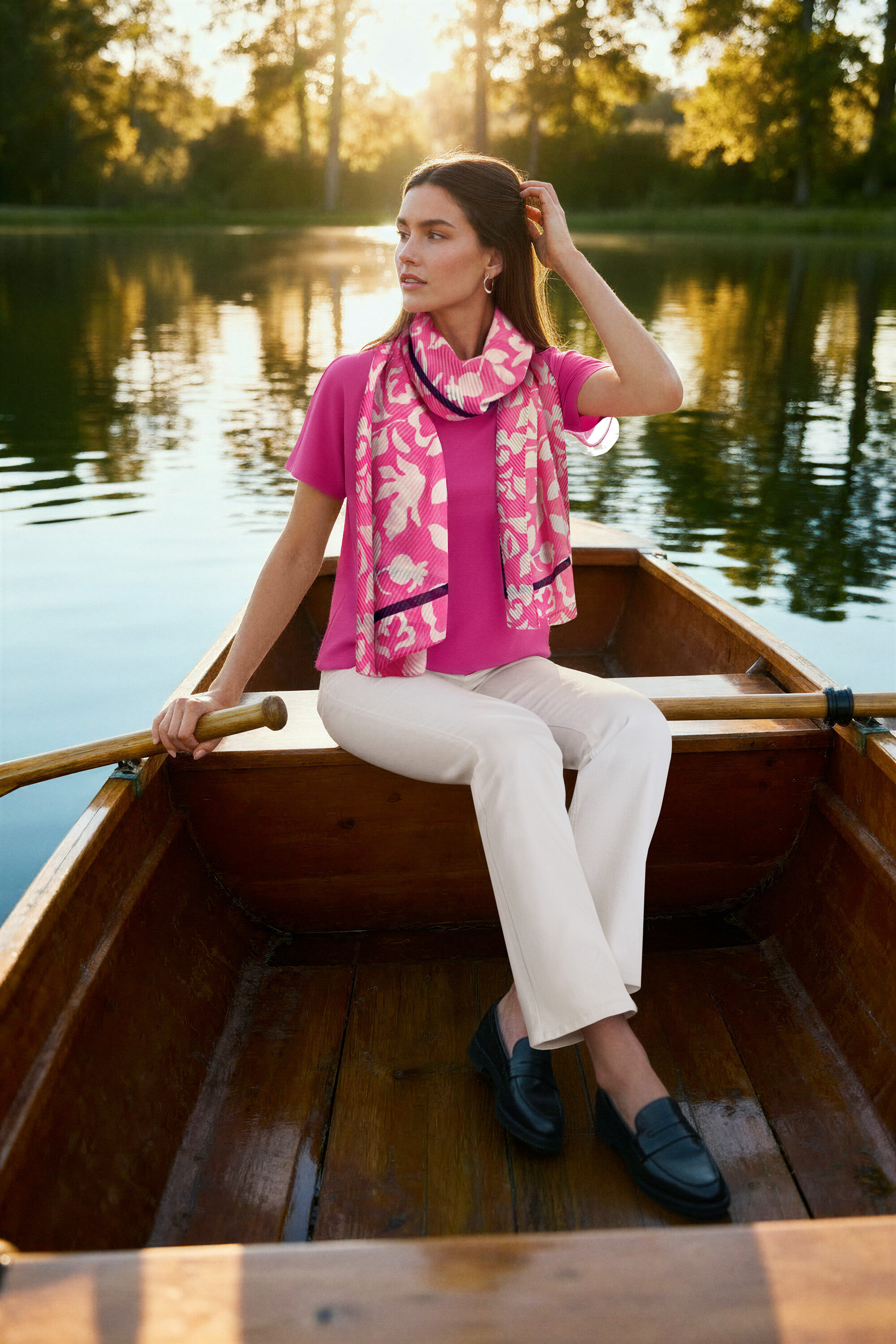 Bir kadın sandalda oturuyor, pembe bir bluz, krem beyazı Bootcut Jean ‒ Fit »Lou«, deri loafer ve Dalga Halka Küpe takıyor.