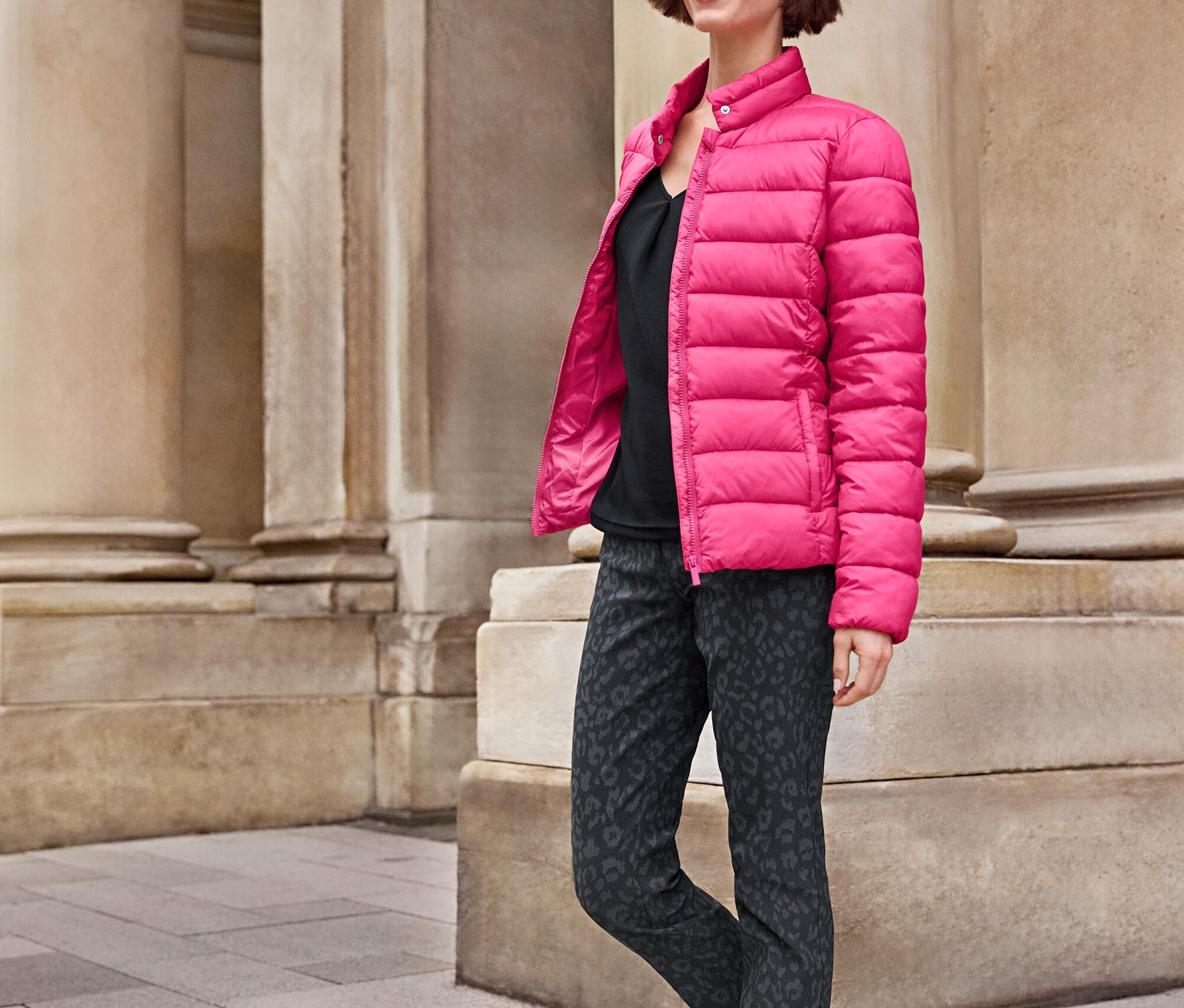 Kadın sütunların yanında yürüyor, pembe bir ceket, desenli pantolon ve beyaz Tom Tailor Sneaker, PU weiß ve 925 Silber Creolen Hexagon giyiyor.