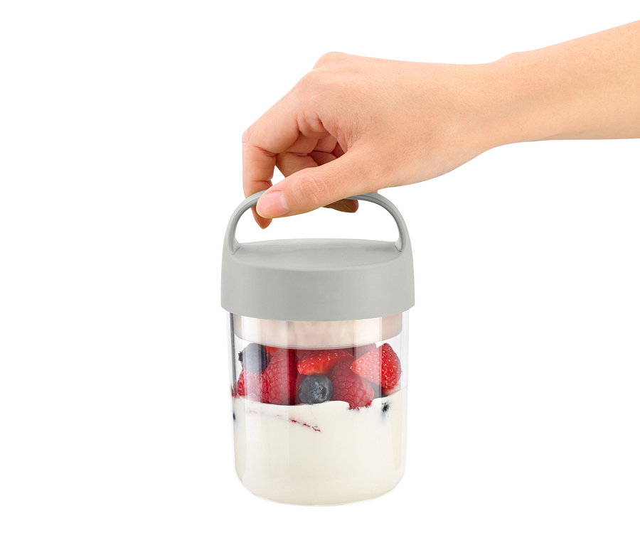Bir el, yoğurt ve meyvelerle dolu taşınabilir bir atıştırmalık kutusu tutuyor.