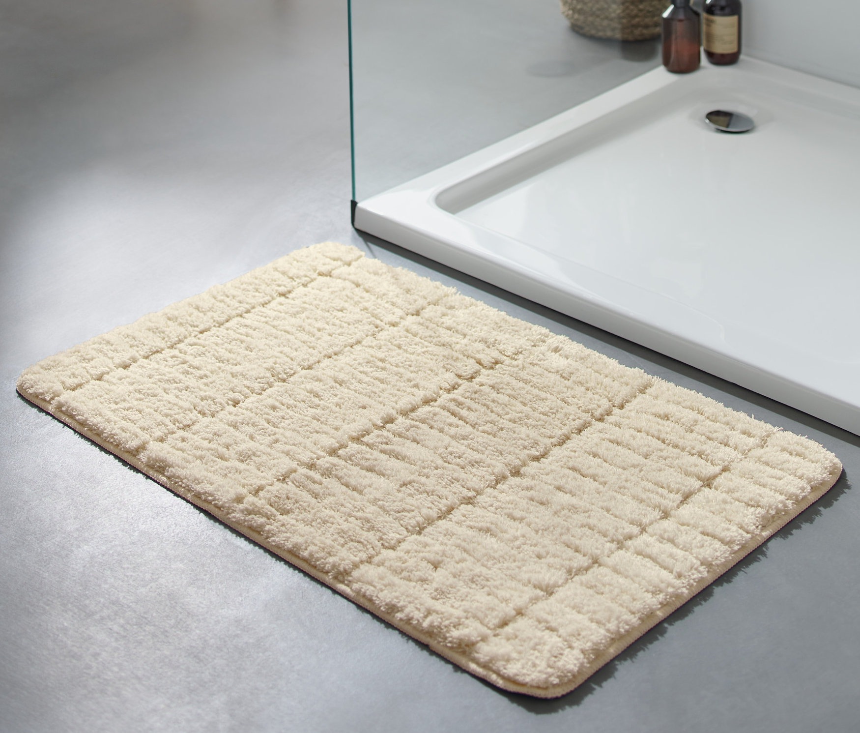 Kırık beyaz renkli, yaklaşık 50 x 80 cm boyutlarındaki Konforlu Banyo Paspası, gri bir zeminde duşakabinin yanında duruyor.