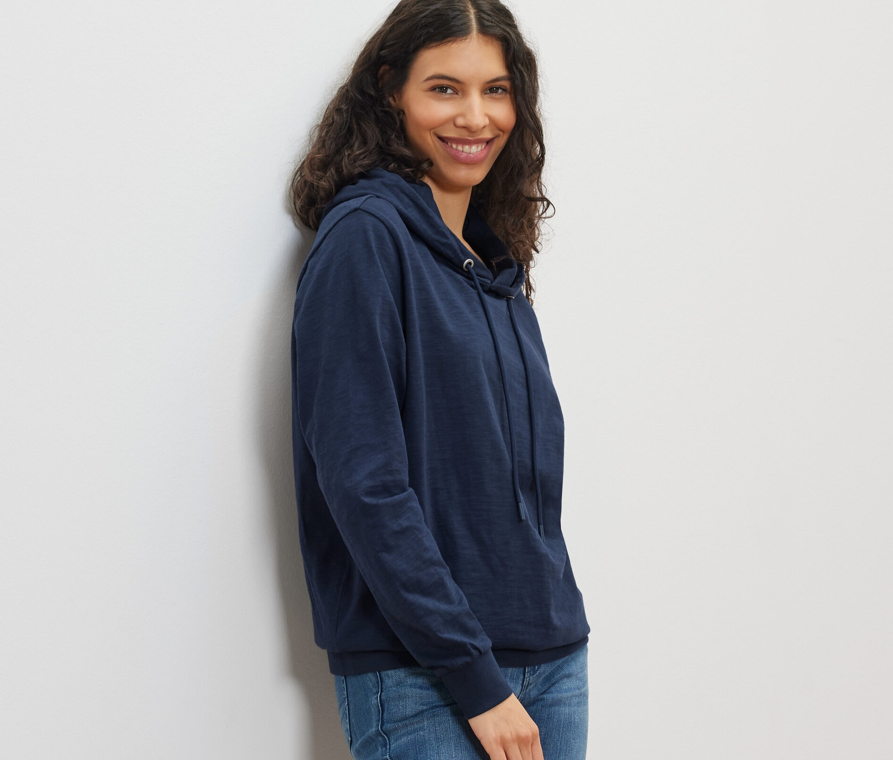 Lacivert jarse kapüşonlu ve kot pantolonlu gülümseyen bir kadın poz veriyor.