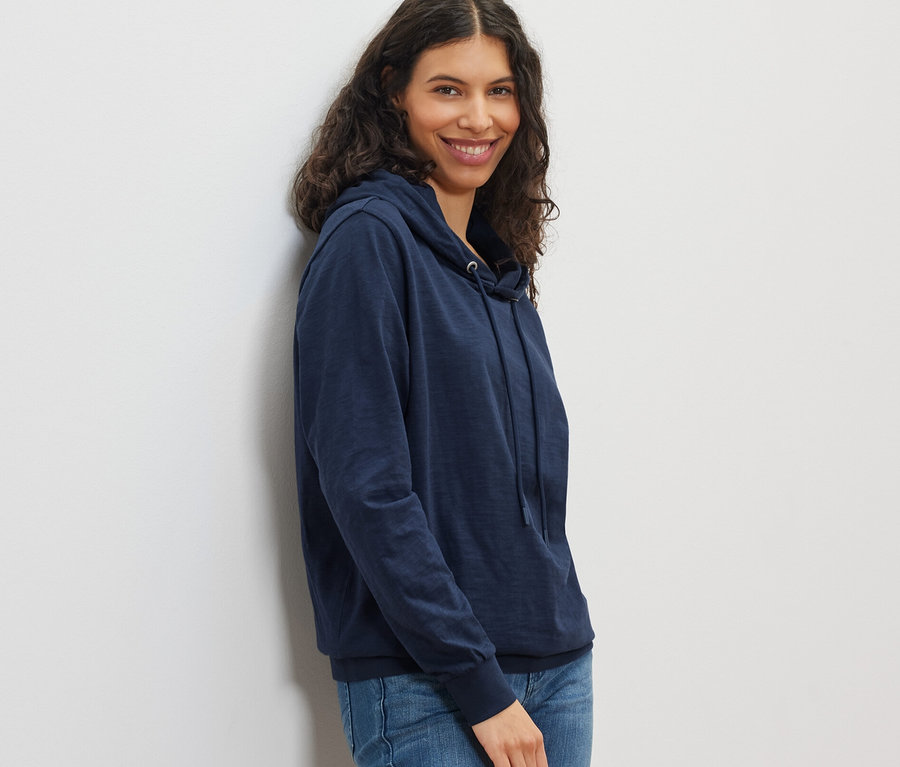 Lacivert jarse kapüşonlu ve kot pantolonlu gülümseyen bir kadın poz veriyor.