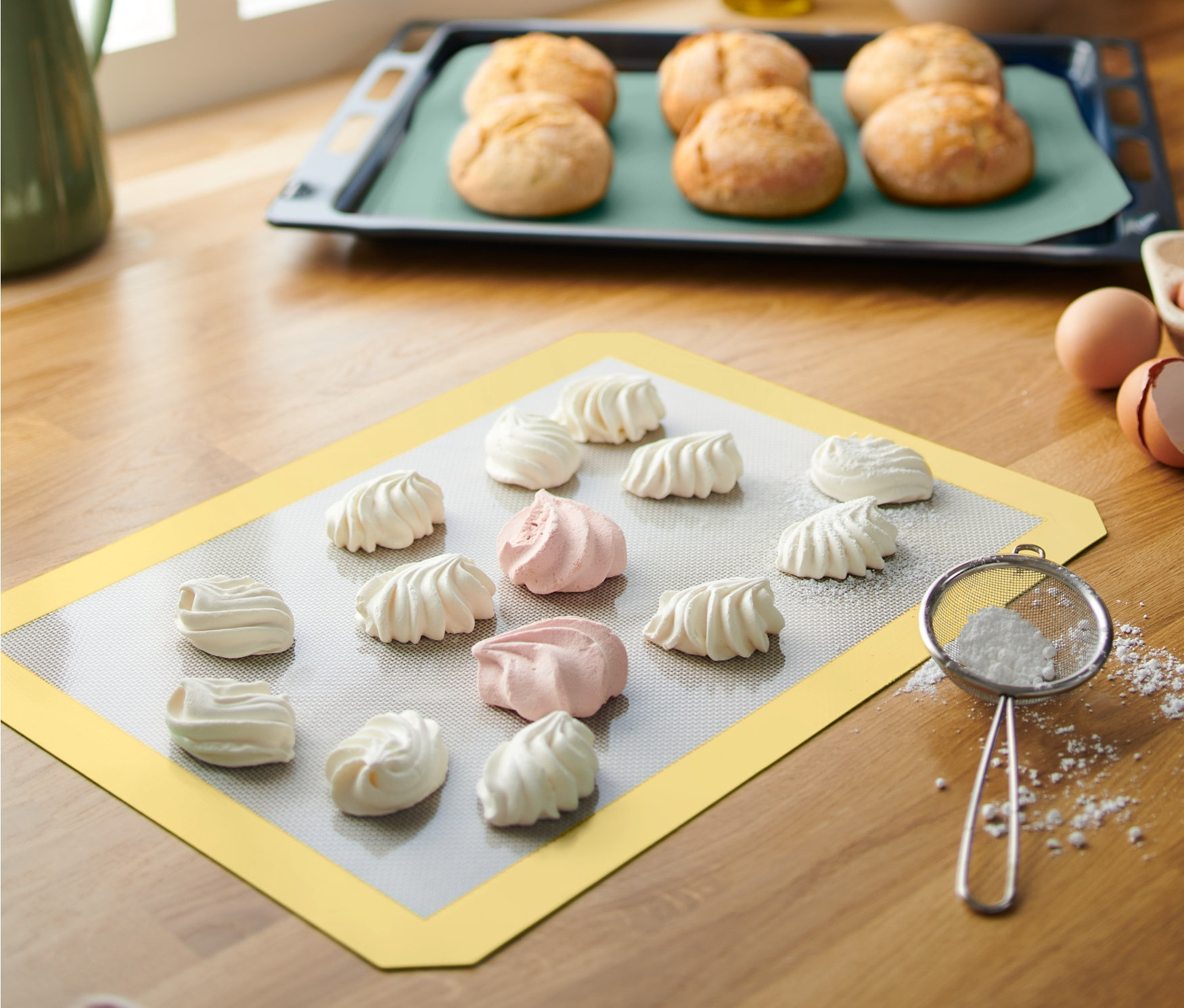 Sarı renkli 2 Adet Profesyonel Dayanıklı Pişirme Matı üzerinde beyaz ve pembe beze kurabiyeler duruyor. Arka planda, bir fırın tepsisi içinde ekmekler var.