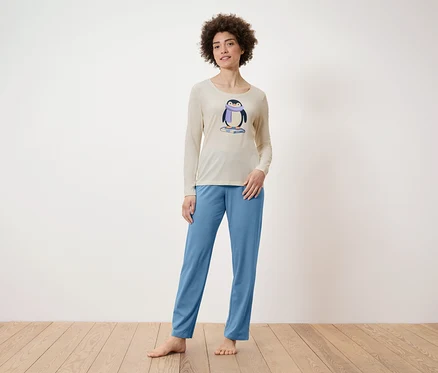 Kadın ayakta duruyor, üzerinde penguen baskılı bej bir pijama üstü ve mavi bir pijama altı var.