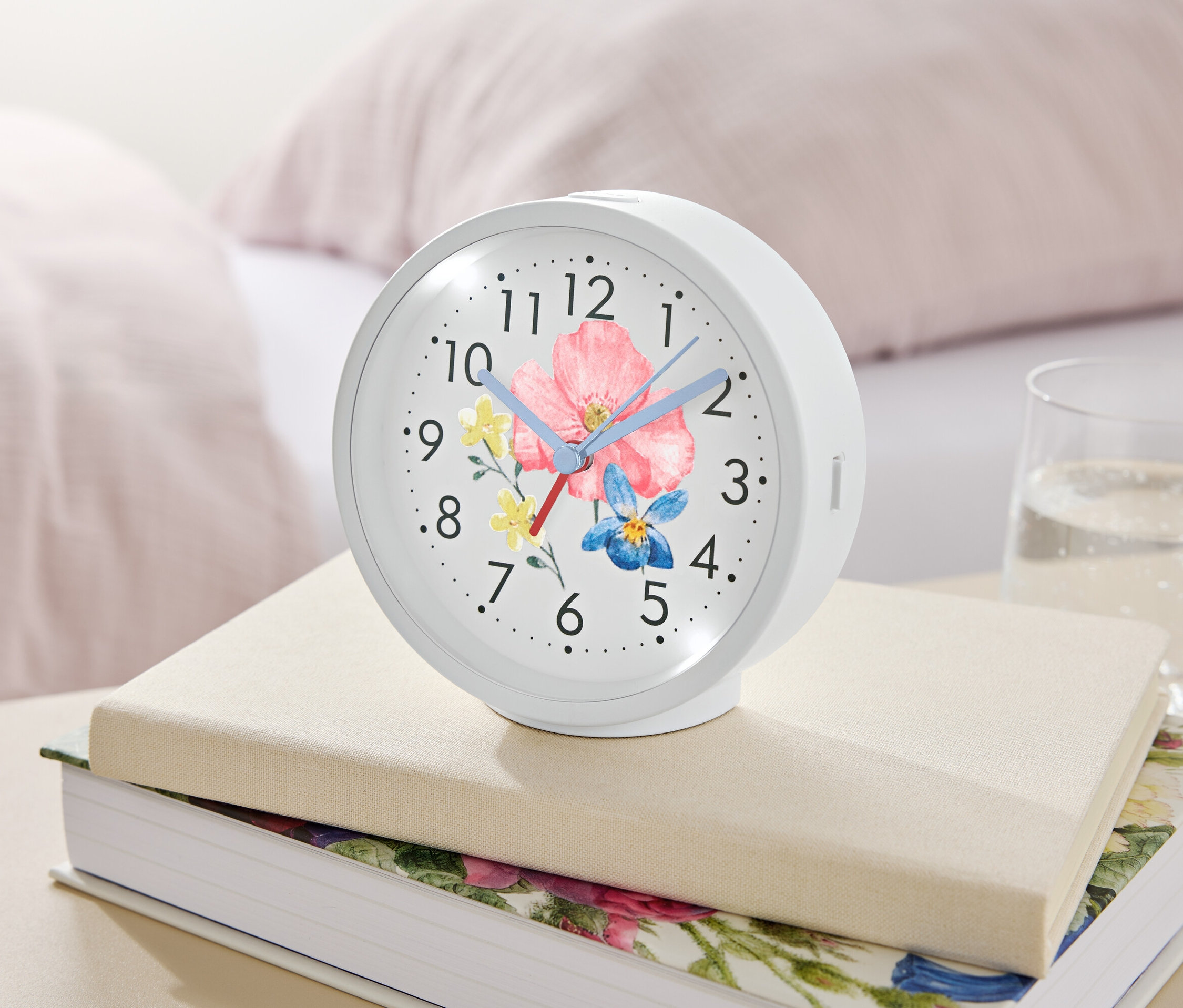 Çiçek desenli beyaz bir çalar saat, bir bardak suyun yanındaki kitapların üzerinde duruyor.