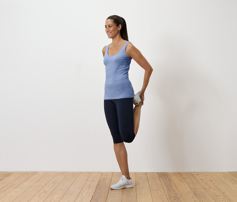 Mavi atlet ve koyu renk tayt giyen kadın, ayağını arkasında tutarak kuadrisepsini esnetiyor.