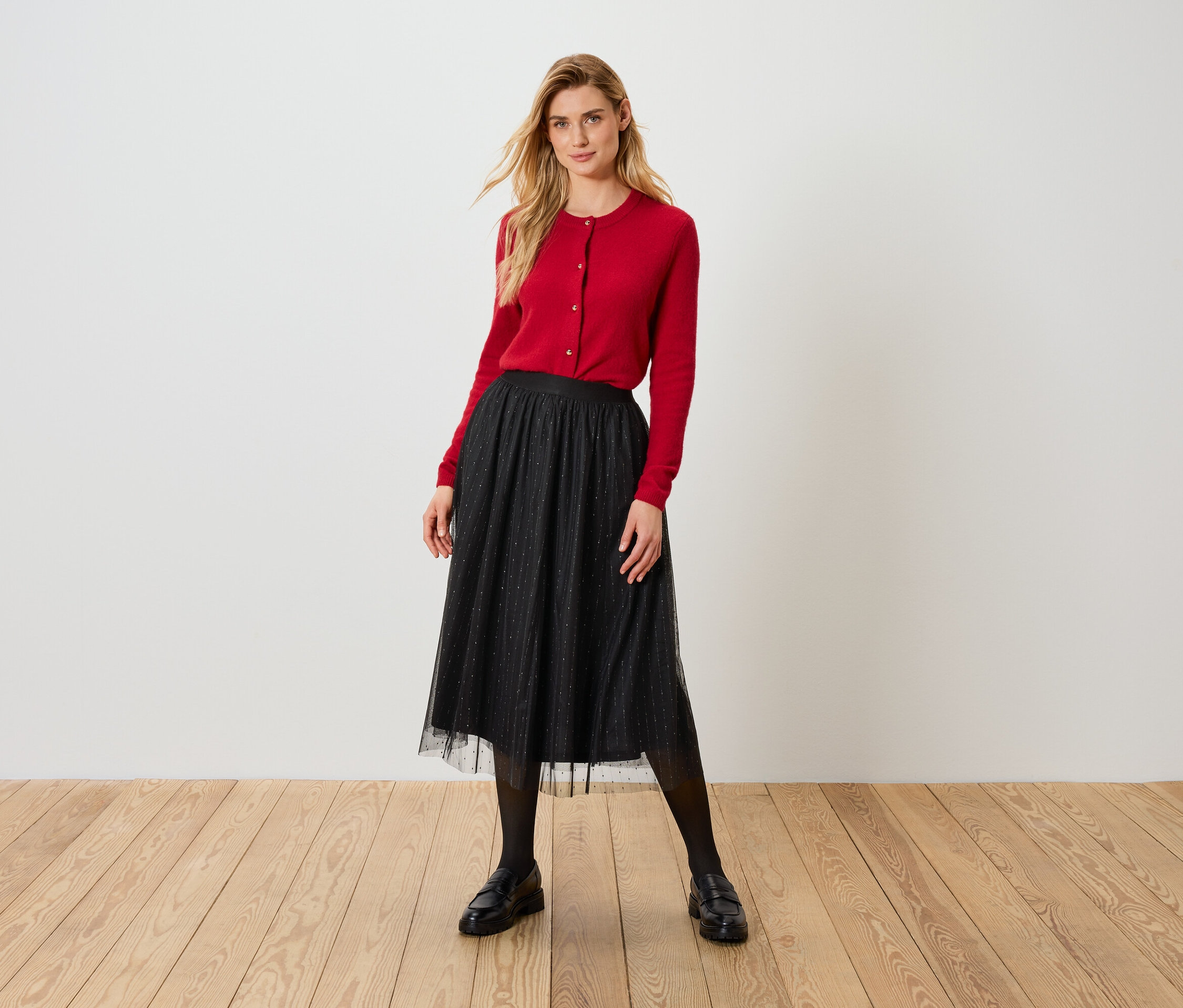 Sarışın kadın kırmızı hırka ve siyah tül etekle ahşap zeminde duruyor.