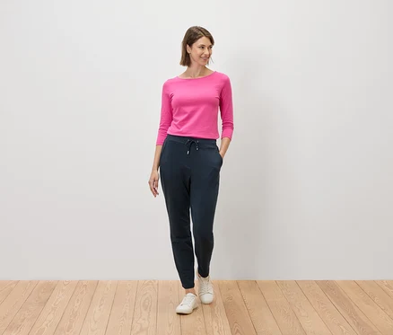 Pembe bluzlu ve lacivert pantolonlu bir kadın, beyaz bir duvarın önünde ahşap bir zeminde duruyor.
