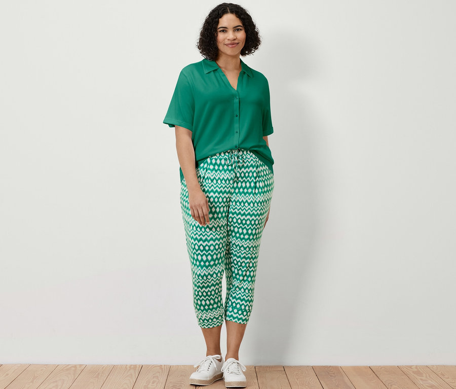 Bir kadın ayakta duruyor, üzerinde zümrüt yeşili düğmeli bluz ve baskılı 3/4 jersey pantolon var.
