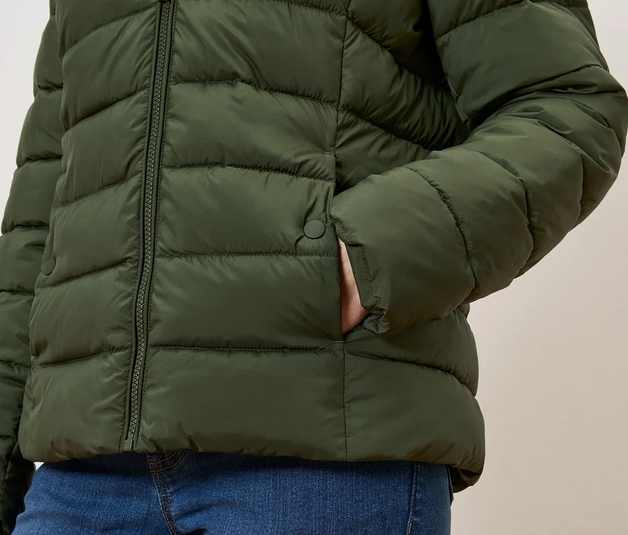 Zeytin yeşili şişme montunun cebinde eli olan, mavi kot pantolonlu kişi.