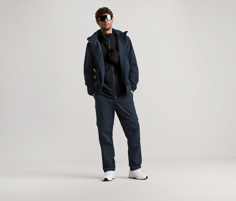 Adam, açık bir arka plan önünde güneş gözlüğü, lacivert ceket, pantolon ve bel çantasıyla duruyor.