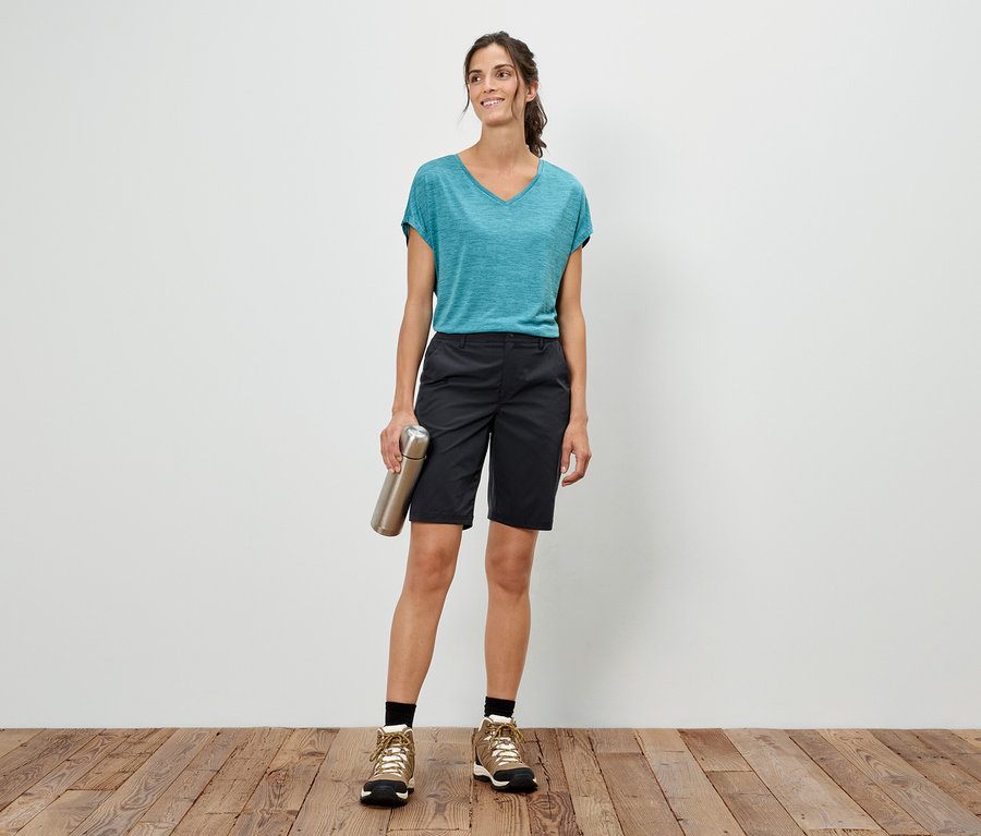 Kadın beyaz bir duvarın önünde duruyor, gümüş bir şişe tutuyor, mavi bir tişört, şort ve yürüyüş ayakkabıları giyiyor.