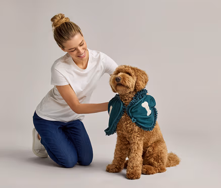 Kadın, kemik aplikeli yeşil pelerin takmış kıvırcık tüylü bir köpeğin yanında diz çökmüş.