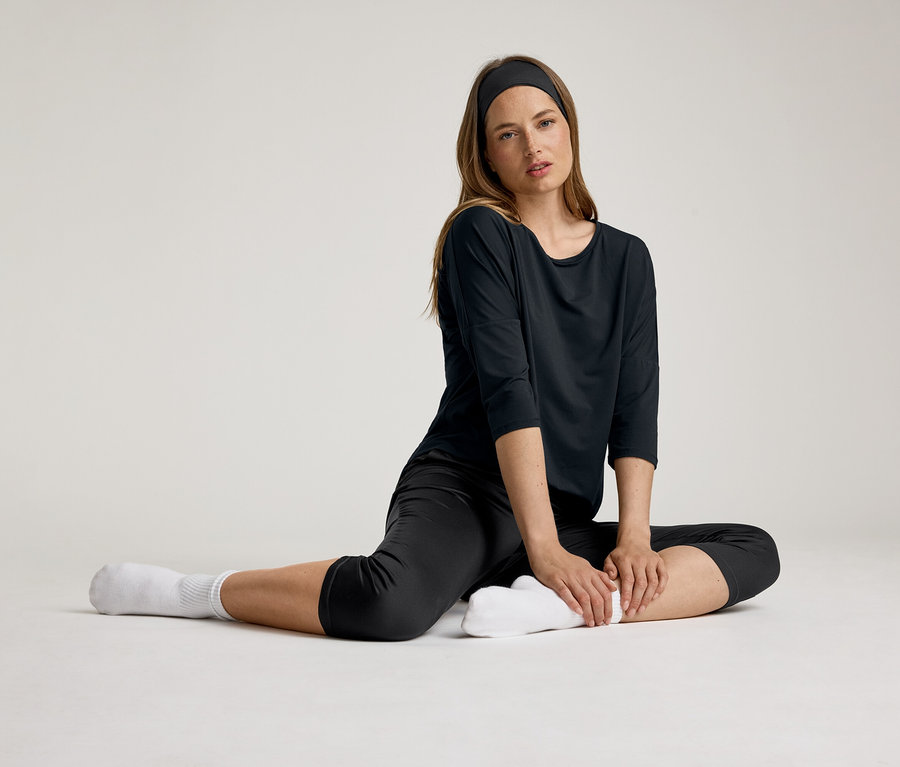 Kadın siyah 3/4 spor tişörtüyle oturuyor. Bir saç bandı ve beyaz çoraplar giyiyor.