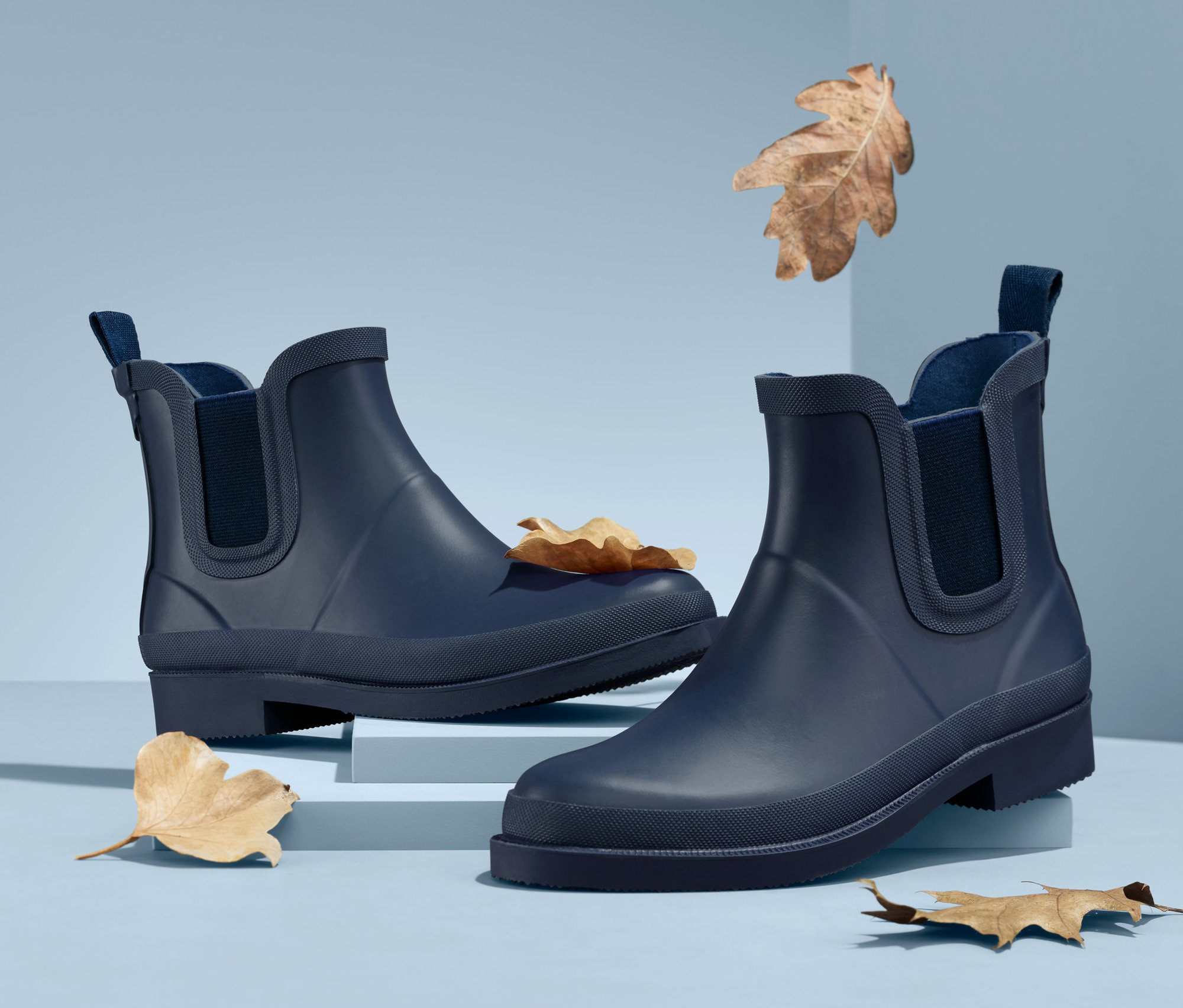 Açık mavi bir arka plan üzerinde iki adet lacivert Yağmur Botu ve kahverengi yapraklar.