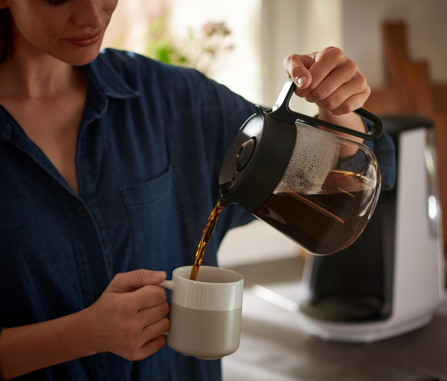 Kadın, bir demlikten bir fincana kahve dolduruyor. Arka planda »Let's Brew« Filtre Kahve Makinesi görülüyor.