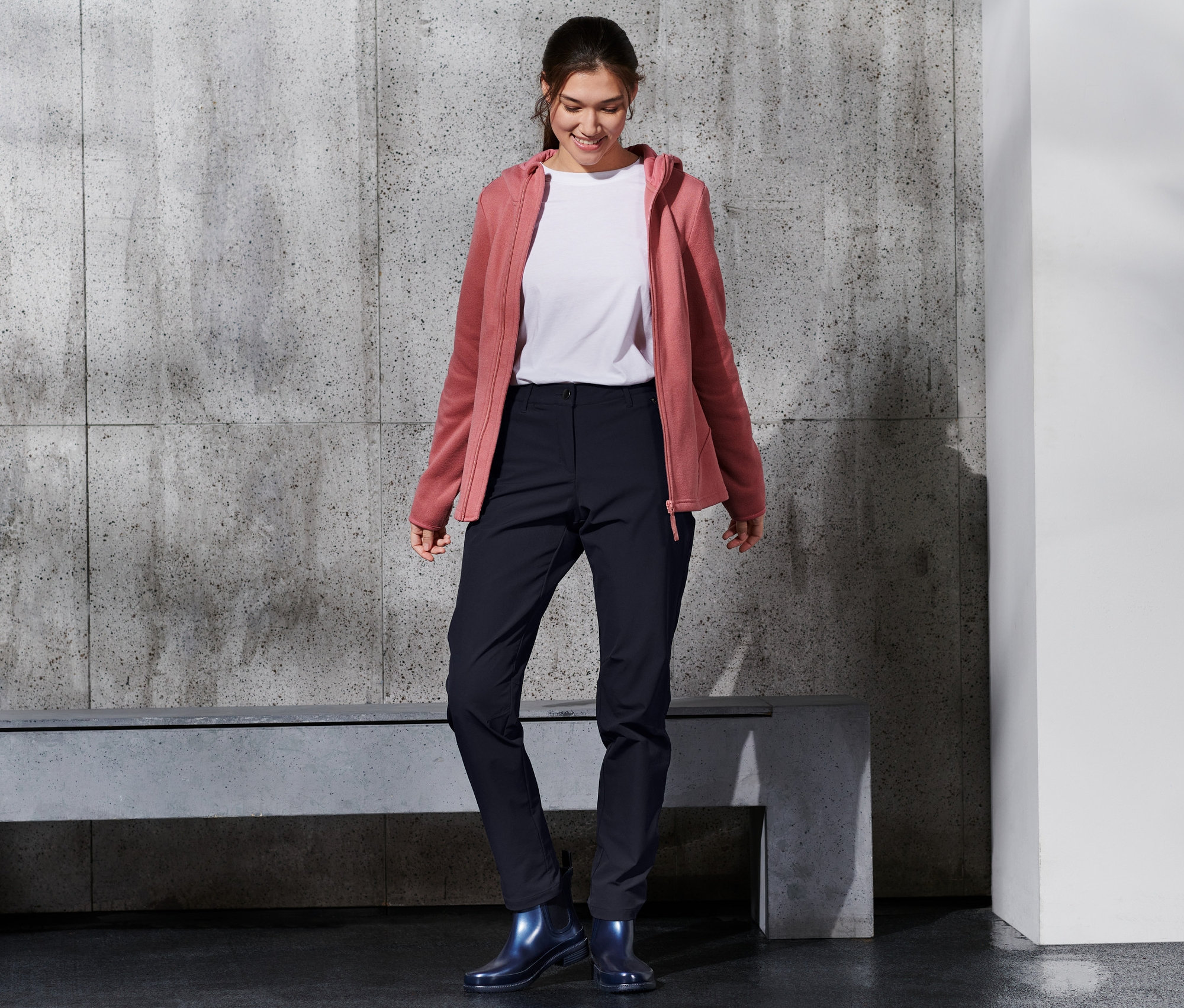 Gülümseyen bir kadın softshell pantolon, beyaz tişört ve pembe kapüşonlu giyiyor.