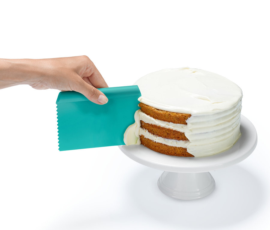 Bir el, Profesyonel Pişirme Yardımcısı Seti'nden turkuaz bir spatula ile üç katlı bir kekin üzerine krema sürüyor.