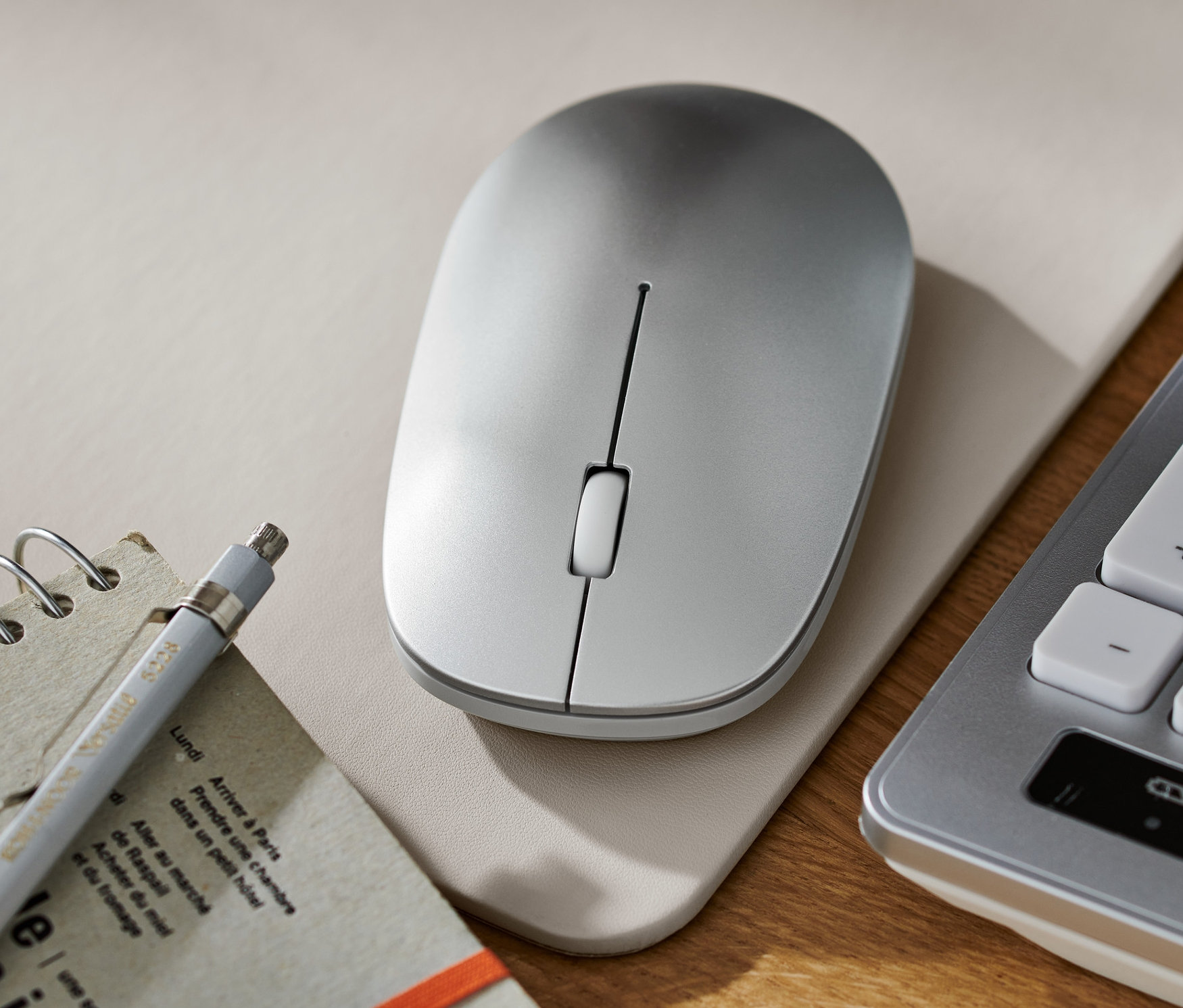 Gümüş kablosuz fare, bir not defteri, kalem ve klavyenin yanında bir ped üzerinde duruyor.