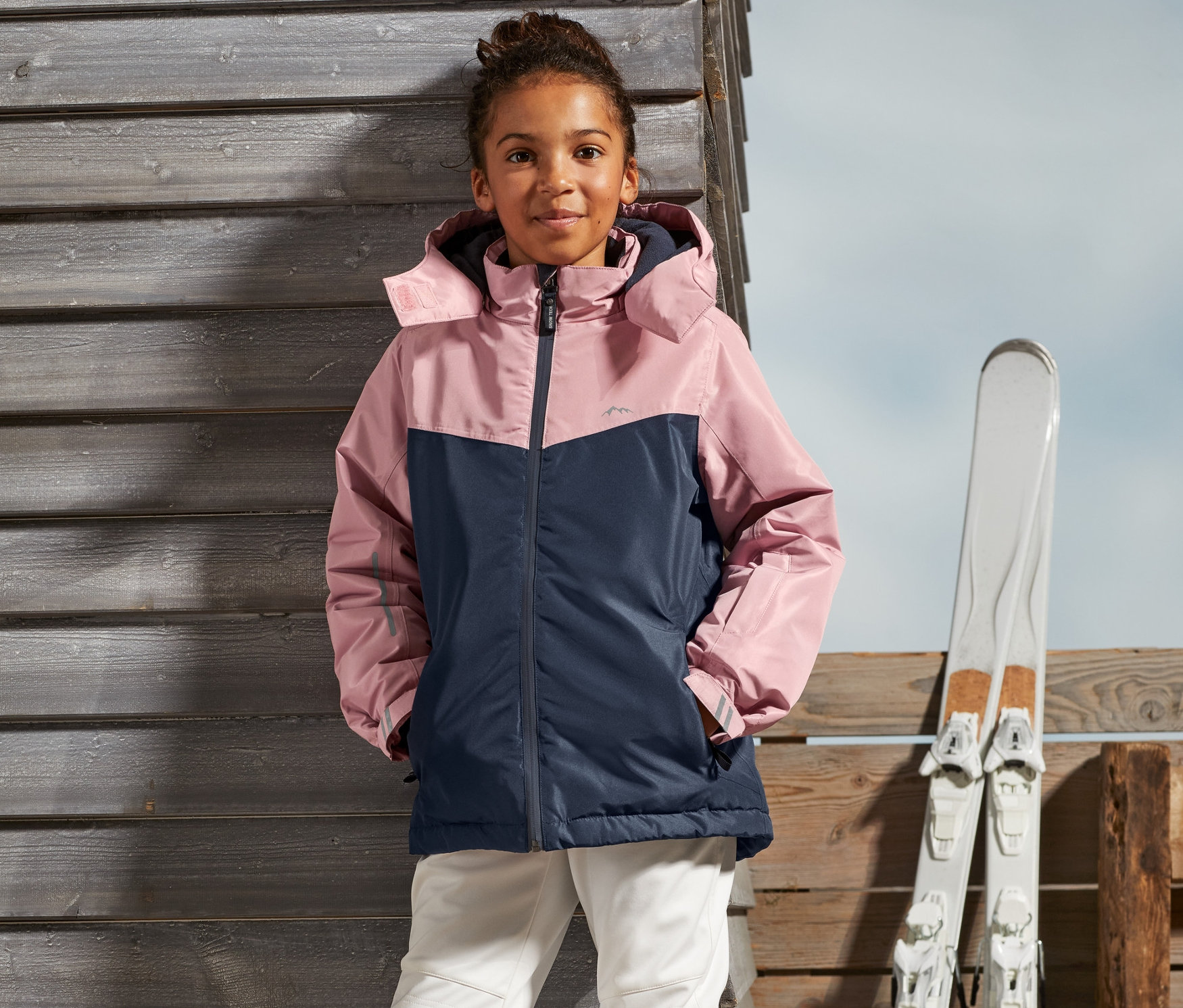 Kız, Kız Çocuk Kayak Montu ve kayak pantolonuyla dışarıda duruyor.