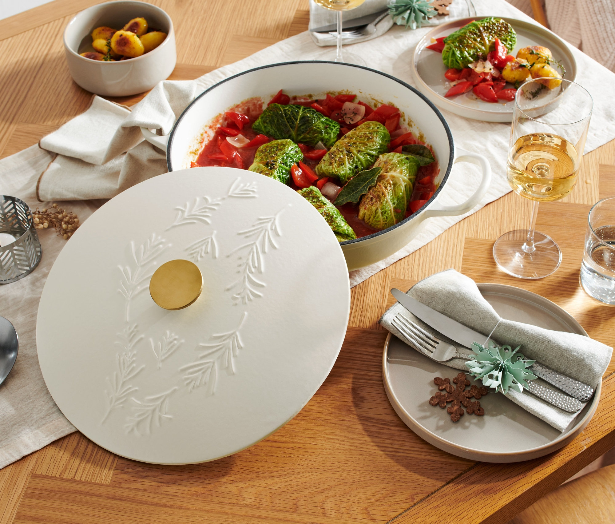 Ahşap bir masanın üzerinde, kabartmalı dal deseni olan kapaklı, beyaz renkli dökme demir bir servis tavası duruyor.