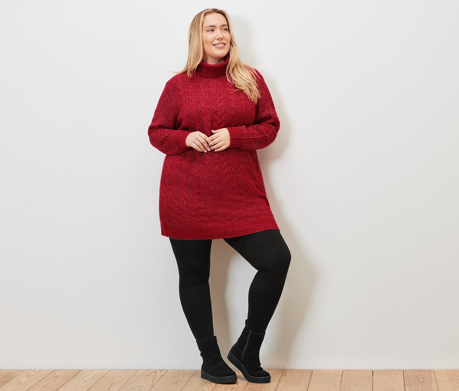 Kadın uzun, kırmızı, saç örgüsü desenli triko kazak, siyah tayt ve botlarla poz veriyor.