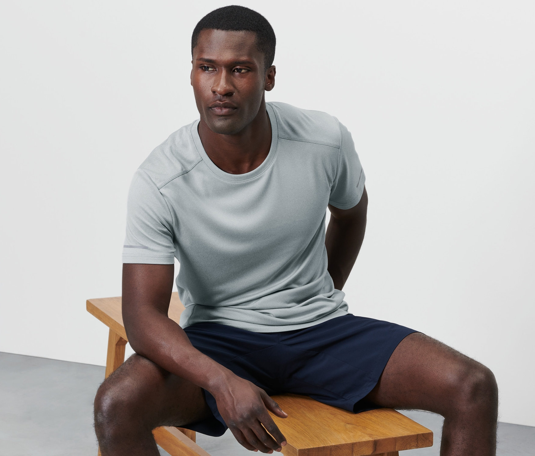 Adam ahşap bir bankta oturuyor ve kırçıllı gri bir fonksiyonel tişört giyiyor.