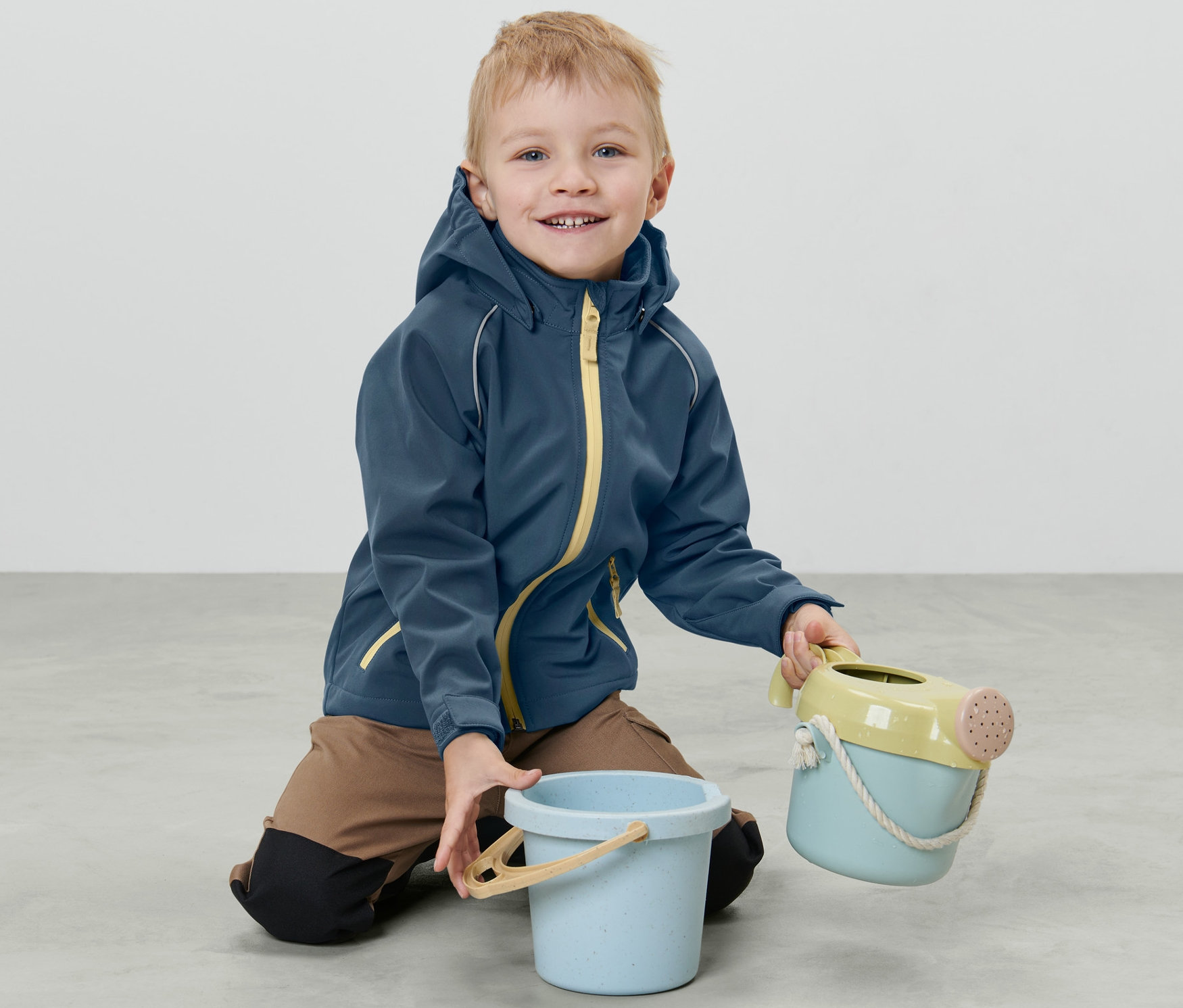 Bir çocuk yerde diz çökmüş, Küçük Çocuk Softshell Ceket giymiş ve oyuncak tutuyor.