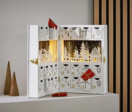 Açık, beyaz renkli, süslemeli ve hediyeli bir advent takvimi, üç adet ahşap koninin yanında duruyor.