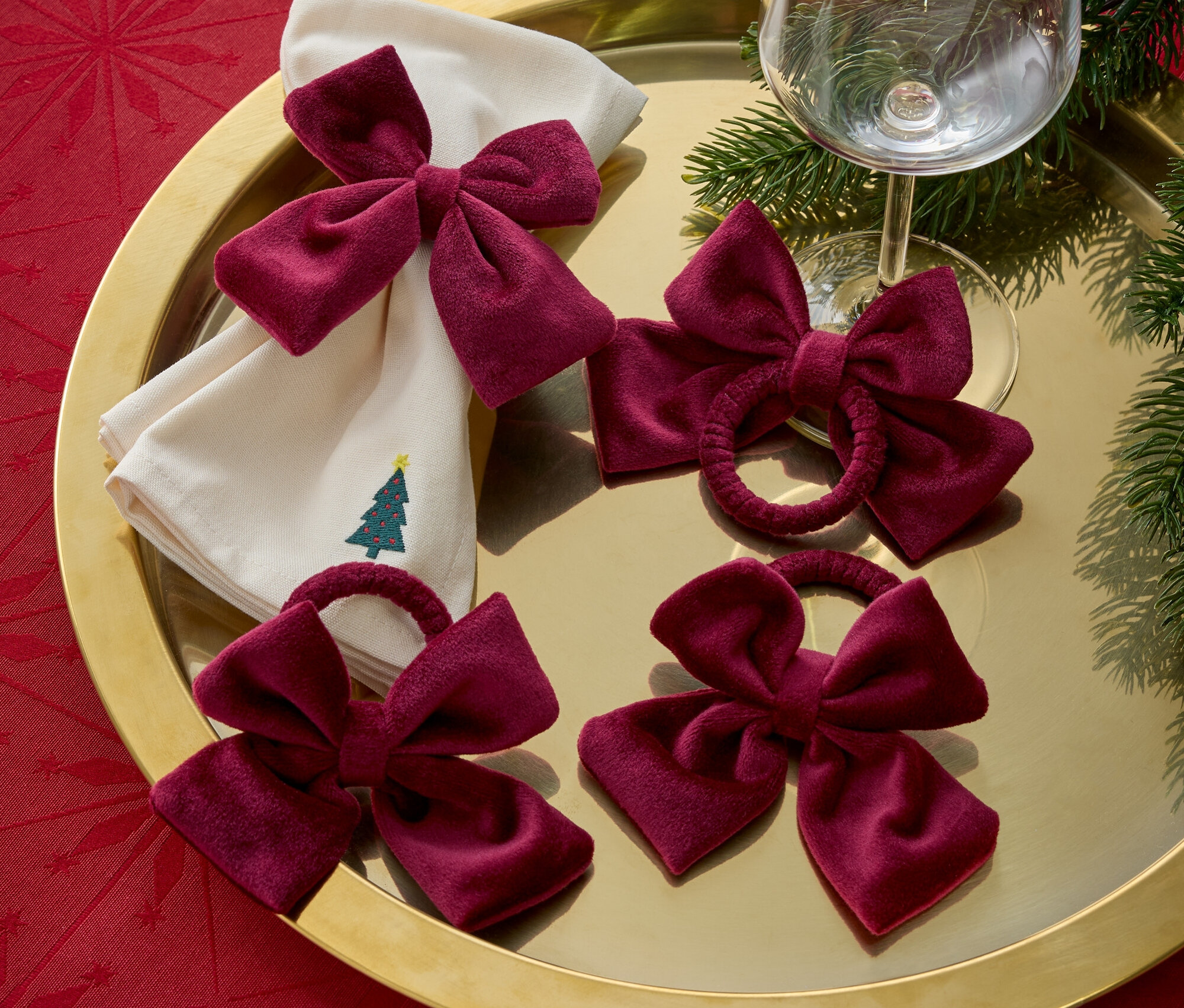 Altın bir tepside Noel ağacı işlemeli kumaş peçeteler, bordo kurdeleler ve bir şarap kadehi bulunmaktadır.