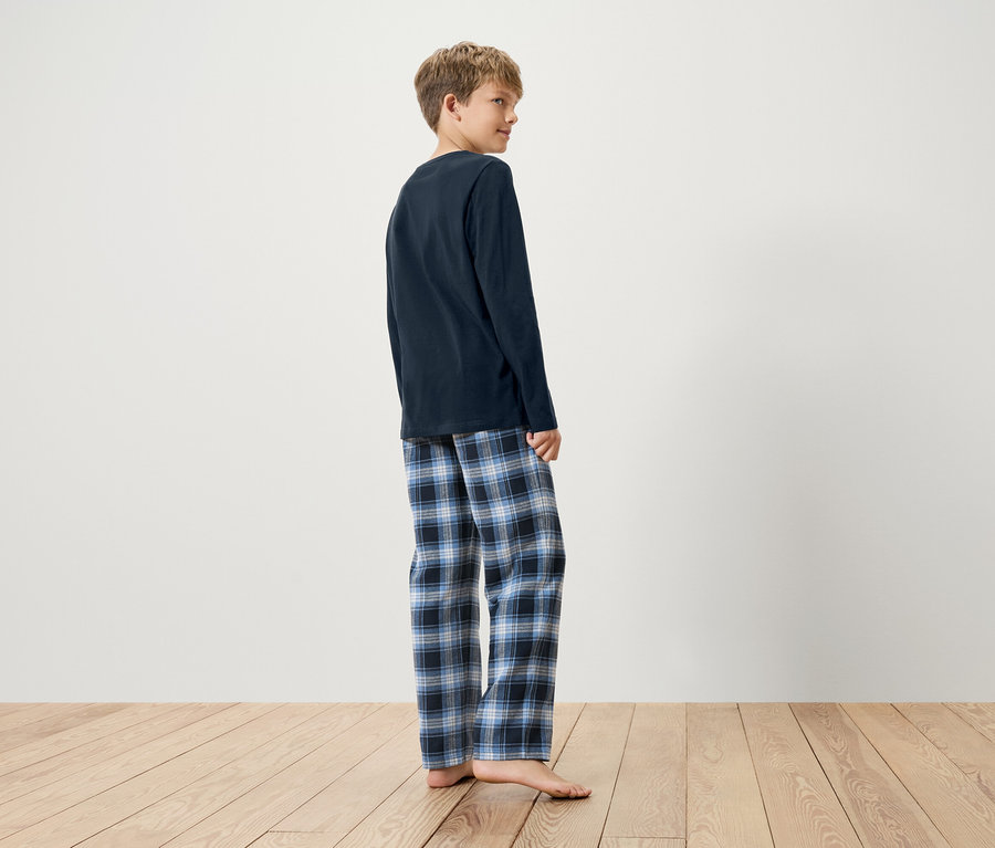 Lacivert uzun kollu pijama üstü ve mavi kareli pijama altı giyen bir çocuk, ahşap zeminde yalın ayak duruyor.