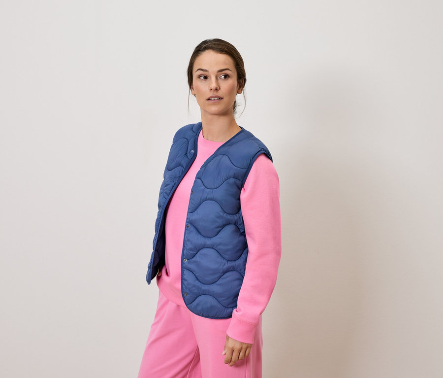 Pembe eşofmanlı ve mavi kapitone yelekli bir kadın ayakta duruyor ve uzaklara bakıyor.