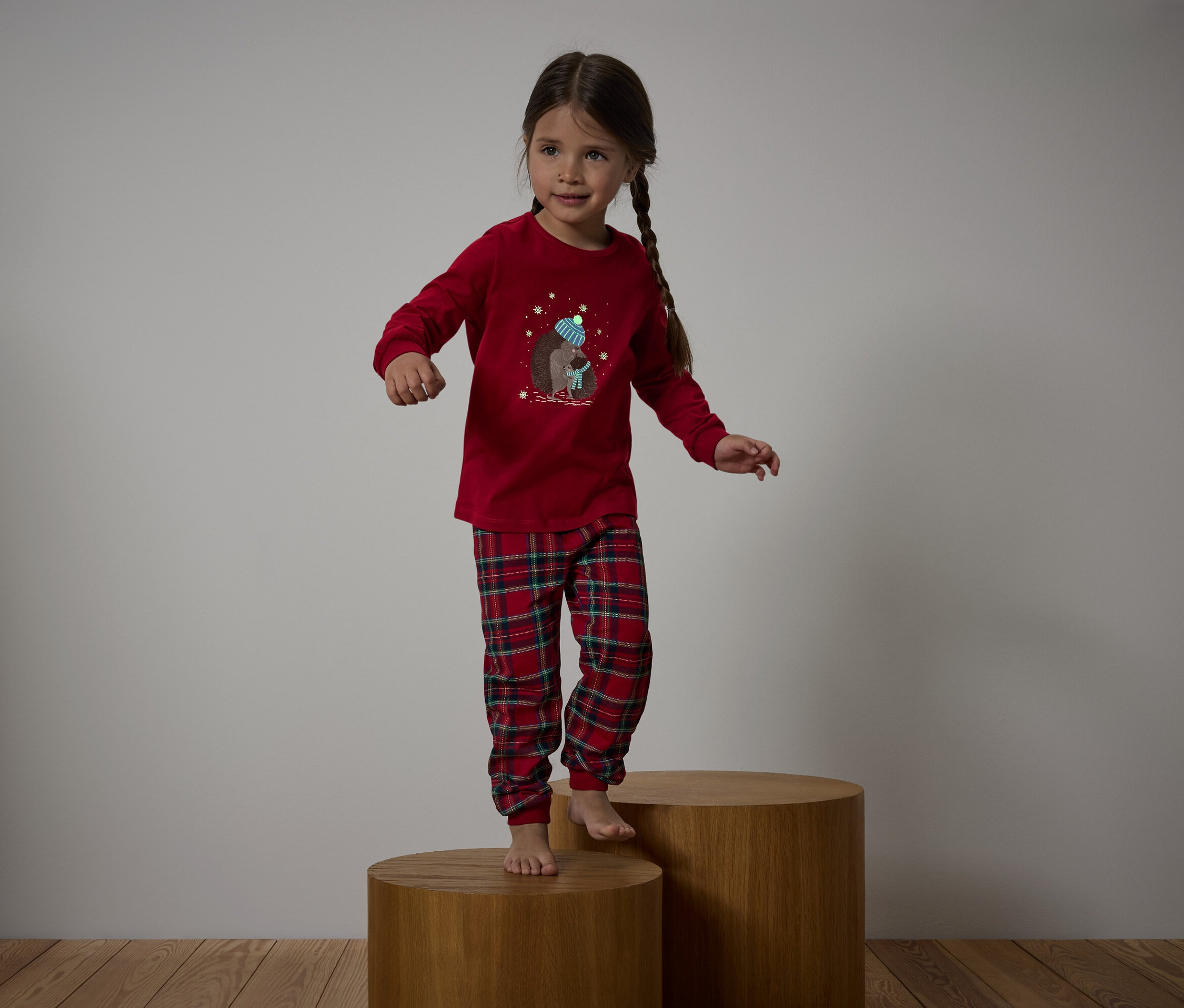 Kareli pantolonlu kırmızı pijamalı bir kız, ahşap kaideler üzerinde duruyor.