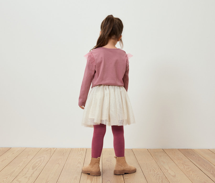 Kameraya arkası dönük kız, pembe bir kazak, parıltılı bir etek, pembe tayt ve kahverengi botlar giyiyor.