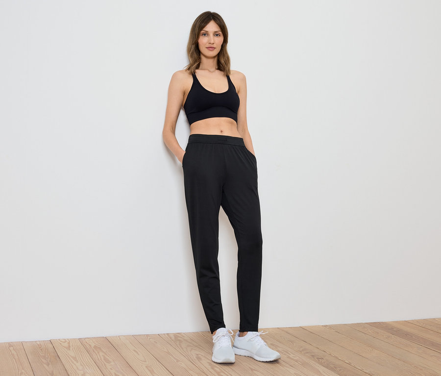 Siyah sporcu sütyeni ve pantolonu giyen bir kadın beyaz bir duvara yaslanmış duruyor.