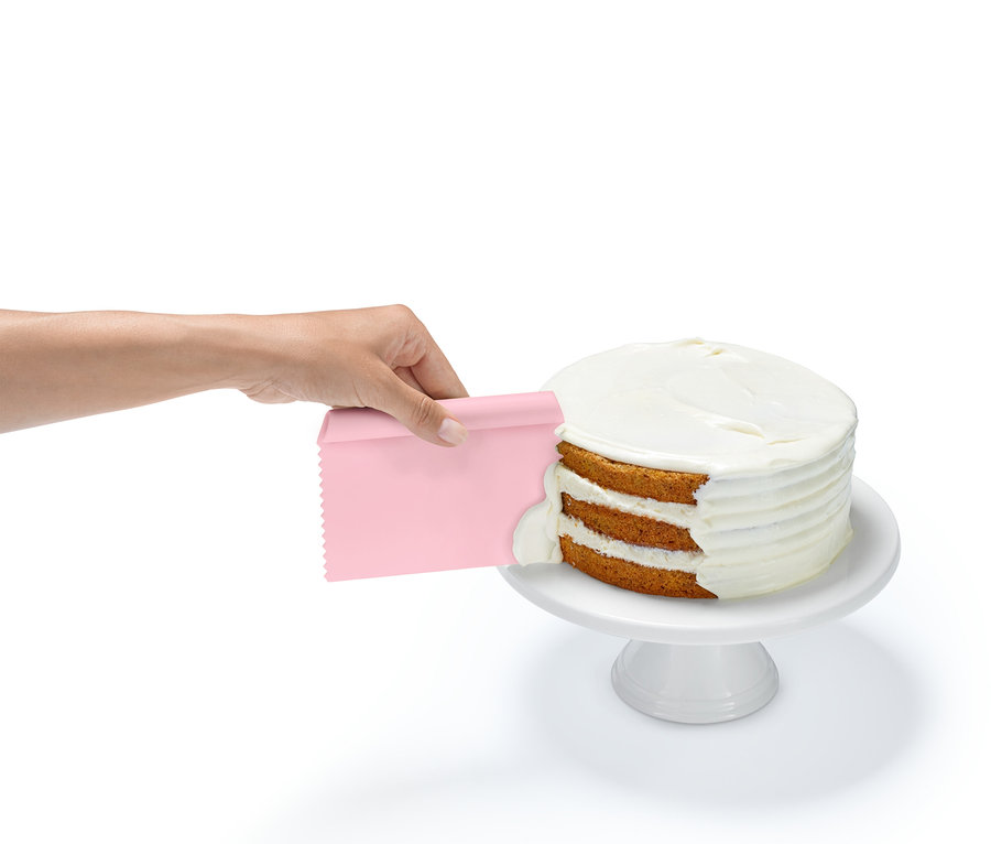 Bir el, beyaz bir taban üzerinde duran üç katlı bir pastaya pembe bir spatula ile krema sürüyor.