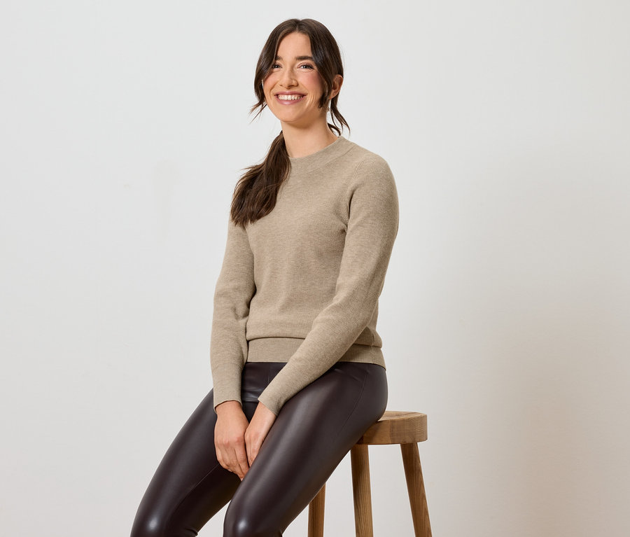 Kahverengi deri tayt ve bej kazak giyen bir kadın taburede oturuyor.