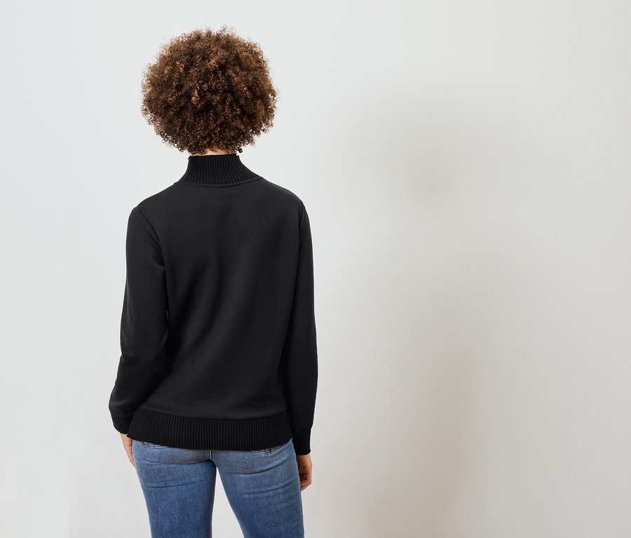 Kıvırcık saçlı, siyah balıkçı yaka ve mavi kot pantolon giyen bir kadının arka görünümü.