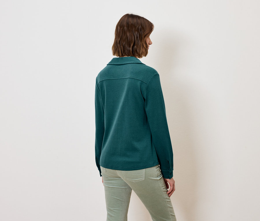 Sırtı kameraya dönük bir kadın, uzun kollu yeşil bir gömlek ve yeşil pantolon giyiyor.