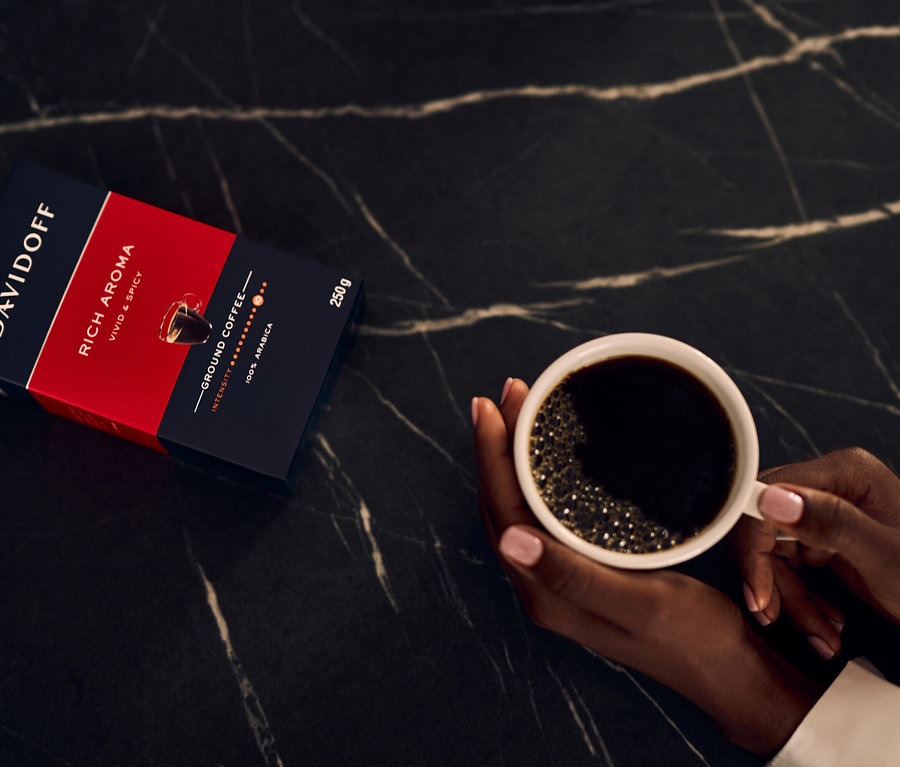 Bir kişi, DAVIDOFF CAFÉ Rich Aroma Gemahlen paketinin yanında bir fincan kahve tutuyor.