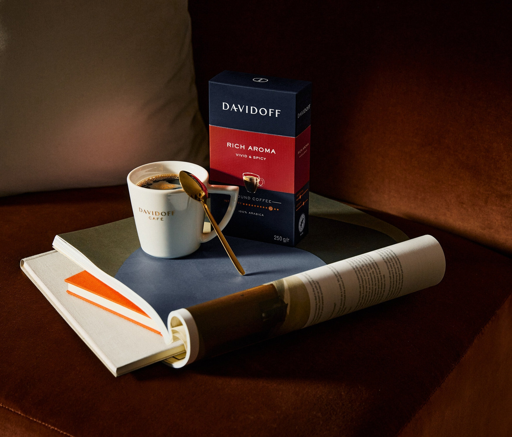 Kahverengi bir kanepede Davidoff Cafe kahve fincanı, Davidoff Rich Aroma paketinin yanında, altın bir kaşık, bir kitap ve rulo yapılmış bir kağıt duruyor.