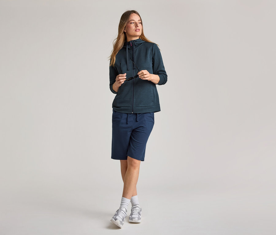 Lacivert kapüşonlu ceket ve şort giyen kadın. Elinde güneş gözlüğü tutuyor.