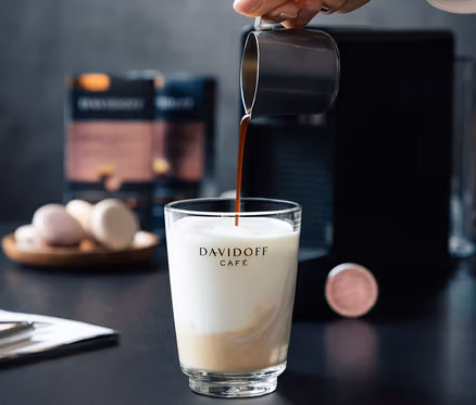 Bir el sütlü bir bardağa kahve döküyor. Arka planda bir kahve makinesi ve bir paket DAVIDOFF CAFÉ Crema Intense bulunuyor.